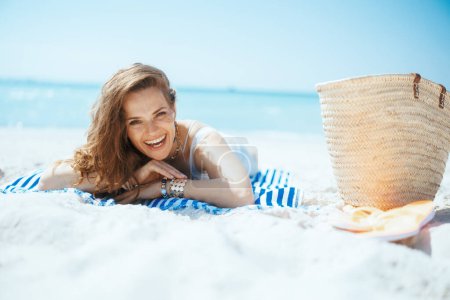 Foto de Sonriente mujer moderna de mediana edad en la costa del océano con bolsa de paja y toalla a rayas. - Imagen libre de derechos