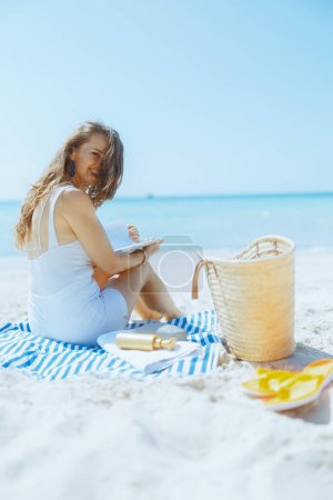 happy elegant middle aged woman on the seashore with straw bag, book and striped towel.