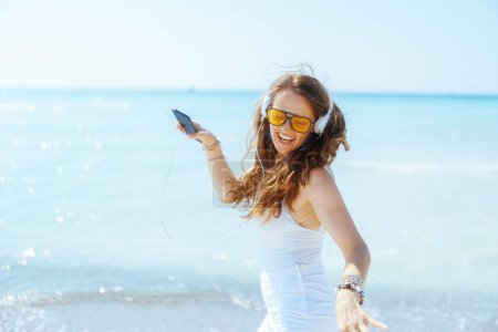 Foto de Sonriente mujer moderna de 40 años en la costa con auriculares y smartphone bailando. - Imagen libre de derechos