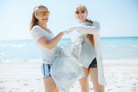 Foto de Mujeres sonrientes voluntarias en la playa con bolsas de basura recogiendo basura. - Imagen libre de derechos