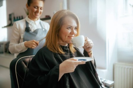 Téléchargez les photos : Coiffeuse dans un salon de beauté moderne avec ciseaux coupe les cheveux et client détendu et tasse de latte de soja. - en image libre de droit