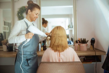 40 Jahre alte Friseurin in modernem Haarstudio mit Haarbürste und Client-Blowout-Haartrockner.