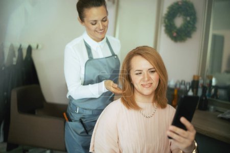 Téléchargez les photos : Femme de 40 ans coiffeuse dans un studio de beauté moderne avec client avec smartphone. - en image libre de droit