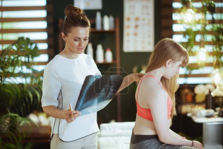 Foto de Tiempo de atención médica. terapeuta de masaje femenino preocupado en gabinete de masaje con rayos X y cliente adolescente haciendo chequeo. - Imagen libre de derechos
