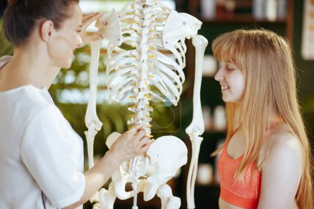 Healthcare time. smiling massage therapist woman in massage cabinet with teenage client and skeleton.