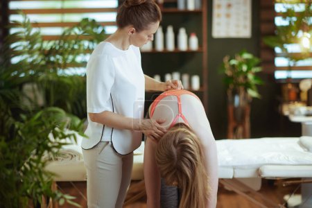 Photo for Healthcare time. pensive female massage therapist in massage cabinet with teenage client checking clients back condition during examination. - Royalty Free Image
