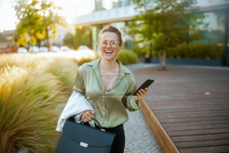 Photo for Smiling modern middle aged woman employee in business district in green blouse and eyeglasses with smartphone and briefcase walking. - Royalty Free Image