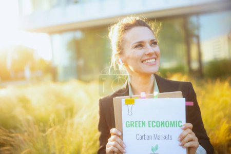 feliz mujer trabajadora moderna cerca del centro de negocios en chaqueta negra con documentos, planes de economía verde, carpeta y auriculares inalámbricos.
