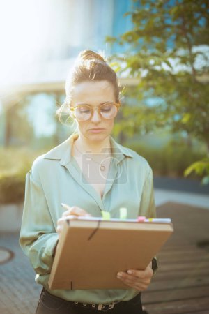 Photo for Pensive modern woman worker near business center in green blouse and eyeglasses with documents and folder. - Royalty Free Image