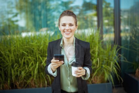 Téléchargez les photos : Heureuse femme d'affaires moderne près du centre d'affaires en veste noire avec smartphone et tasse de café. - en image libre de droit