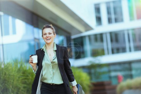 Foto de Sonriente moderna mujer de 40 años empleada cerca del centro de negocios en chaqueta negra con taza de café caminando. - Imagen libre de derechos
