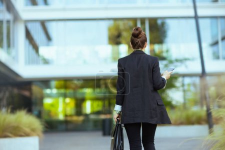 Téléchargez les photos : Vu de derrière femme d'affaires près de l'immeuble de bureaux en veste noire avec mallette en utilisant smartphone et marche. - en image libre de droit