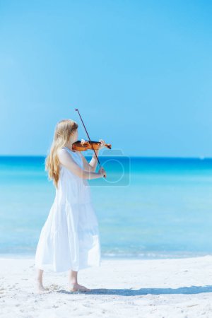 Photo for Full length portrait of modern teenage girl in white dress with violin enjoying playing on the seacoast. - Royalty Free Image