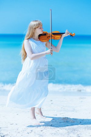 Téléchargez les photos : Portrait complet de jeune femme moderne en robe blanche avec violon jouant sur le bord de la mer. - en image libre de droit