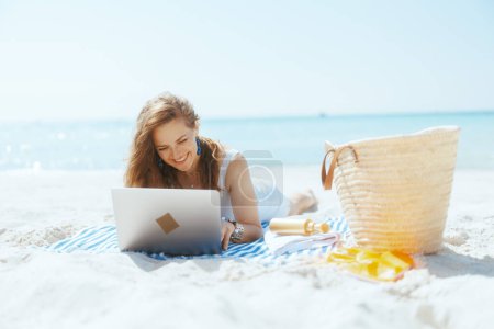 Photo for Smiling modern woman on the ocean coast with straw bag and striped towel using laptop. - Royalty Free Image