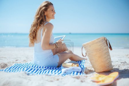 Foto de Mujer feliz y elegante en la playa con bolsa de paja y toalla a rayas utilizando aplicaciones en la tableta PC. - Imagen libre de derechos