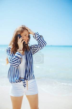 Foto de Sonriente mujer con estilo en la orilla del mar hablando en un teléfono inteligente. - Imagen libre de derechos