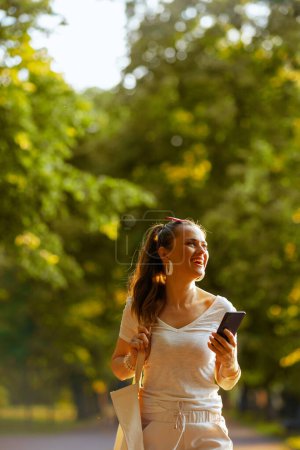 Foto de Hora de verano. mujer joven feliz en camisa blanca con bolso de mano usando teléfono inteligente y caminando en el parque de la ciudad. - Imagen libre de derechos