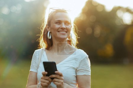 Foto de Hora de verano. sonriente mujer de 40 años de edad en camisa blanca usando aplicación de teléfono inteligente en el prado en el parque de la ciudad. - Imagen libre de derechos