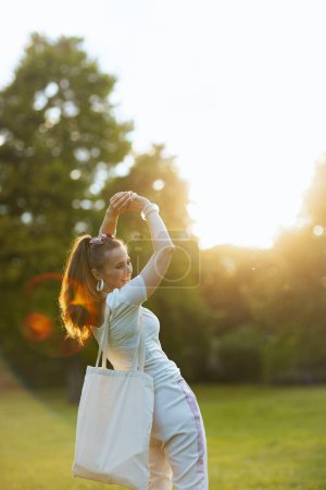 Sommerzeit. fröhliche moderne Frau im weißen Hemd mit Tragetasche auf der Wiese im Stadtpark.