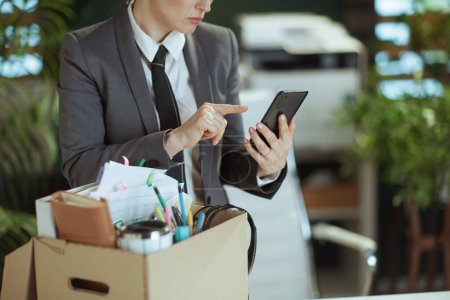 Un nuevo trabajo. infeliz empleada moderna de mediana edad en moderna oficina verde en traje de negocios gris con pertenencias personales en caja de cartón usando teléfono inteligente.