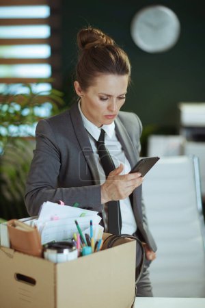 Un nuevo trabajo. infeliz mujer de mediana edad moderna trabajadora en moderna oficina verde en traje de negocios gris con pertenencias personales en caja de cartón y teléfono inteligente.