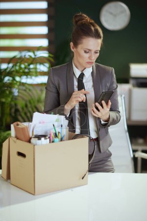 Photo for New job. concerned modern 40 years old woman employee in modern green office in grey business suit with personal belongings in cardboard box speaking on a smartphone. - Royalty Free Image