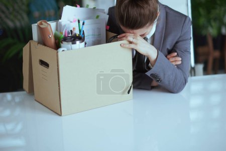 New job. stressed modern female employee in modern green office in grey business suit with personal belongings in cardboard box.