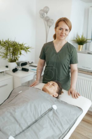 Tiempo de atención médica. terapeuta de masaje médico mujer feliz en el gabinete de masaje con el cliente haciendo procedimiento de envoltura de whisky en manta térmica.