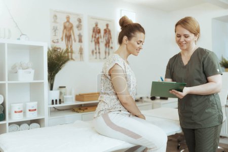 Photo for Healthcare time. smiling massage therapist woman in massage cabinet with client doing checkup and writing in clipboard. - Royalty Free Image