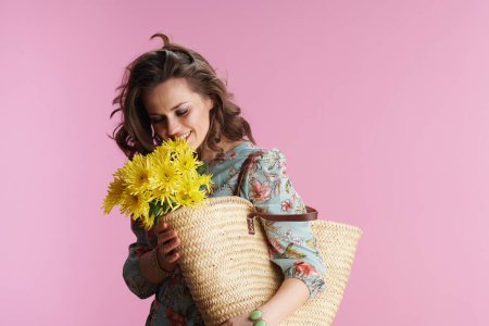 Téléchargez les photos : Femme d'âge moyen souriante avec de longs cheveux bruns ondulés avec des fleurs de chrysanthèmes jaunes et sac de paille sur fond rose. - en image libre de droit