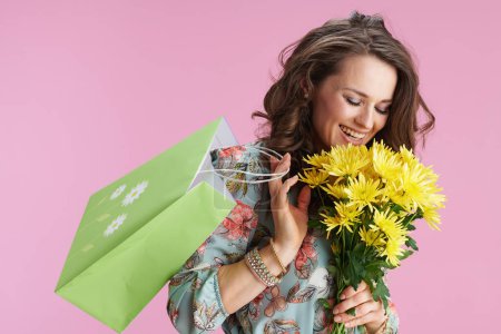 Foto de Mujer moderna feliz con el pelo largo y ondulado morena con flores de crisantemos amarillos y bolsa de compras verde aislado en rosa. - Imagen libre de derechos