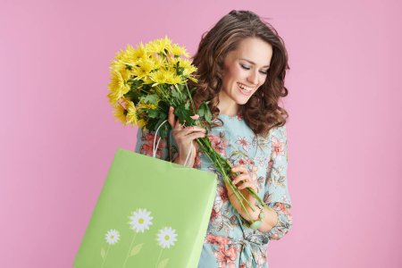 Photo for Smiling stylish woman in floral dress with yellow chrysanthemums flowers and green shopping bag isolated on pink background. - Royalty Free Image