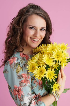 Porträt einer glücklichen stilvollen Frau mit langen welligen brünetten Haaren mit gelben Chrysanthemen-Blumen vor rosa Hintergrund.