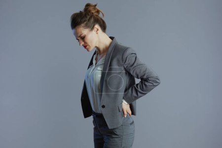 cansada elegante mujer de 40 años empleada en traje gris con dolor de espalda aislado sobre fondo gris.