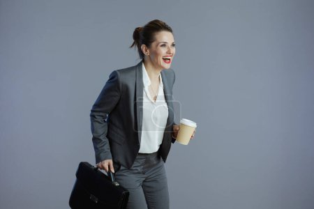 Foto de Sonriente empleada moderna de mediana edad en traje gris con taza de café y maletín corriendo sobre fondo gris. - Imagen libre de derechos