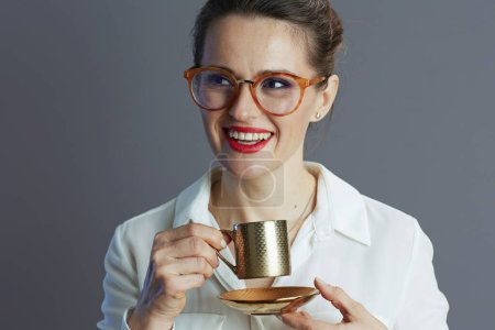 Lächelnde 40-jährige Geschäftsfrau in weißer Bluse mit Gläsern und Kaffeetasse auf grauem Hintergrund.