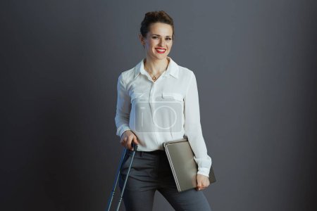 Foto de Sonriente mujer de negocios moderna en blusa blanca con portátil y bolsa de ruedas aislada en gris. - Imagen libre de derechos