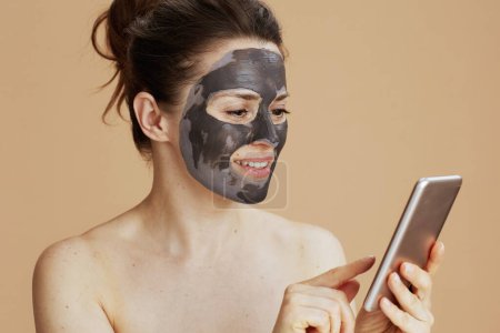Foto de Mujer joven sonriente con máscara facial usando teléfono inteligente contra fondo beige. - Imagen libre de derechos