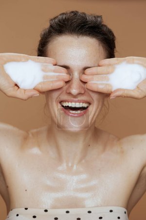 Foto de Mujer de mediana edad feliz con espuma facial limpiador lavado cara aislada sobre fondo beige. - Imagen libre de derechos