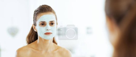 Téléchargez les photos : Jeune femme avec masque cosmétique facial regardant dans le miroir - en image libre de droit