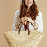 Beach vacation. happy stylish housewife in white blouse and shorts on beige background with straw bag listening to the music with headphones.