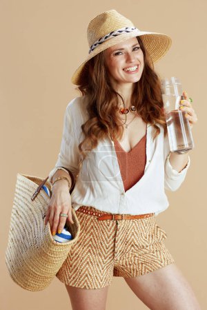 Beach vacation. happy stylish middle aged housewife in white blouse and shorts against beige background with bottle of water, straw bag and summer hat.