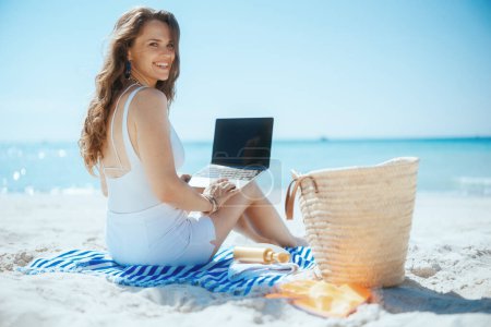 Téléchargez les photos : Heureuse élégante femelle sur la côte de l'océan avec sac de paille, écran blanc d'ordinateur portable et serviette rayée. - en image libre de droit