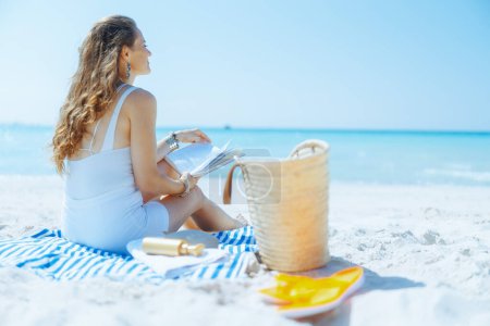 Téléchargez les photos : Femme d'âge moyen moderne détendue sur le littoral avec sac de paille, livre et serviette rayée. - en image libre de droit