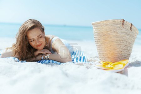 Foto de Relajado elegante mujer de 40 años en la costa con bolsa de paja y toalla a rayas. - Imagen libre de derechos