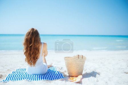 Seen from behind stylish 40 years old woman on the seashore with straw bag and striped towel.