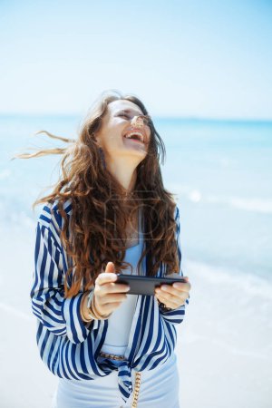 Stilvoll lächelnde 40-jährige Frau mit Smartphone an der Küste.