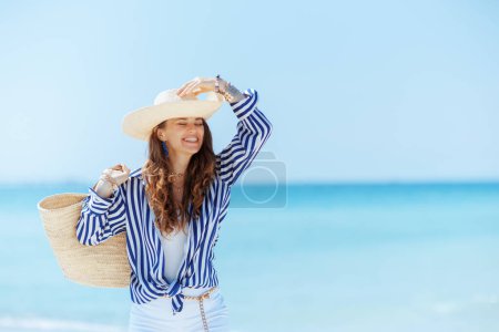 Téléchargez les photos : Heureuse femme d'âge moyen élégant sur la côte de l'océan avec sac de paille et chapeau de paille. - en image libre de droit