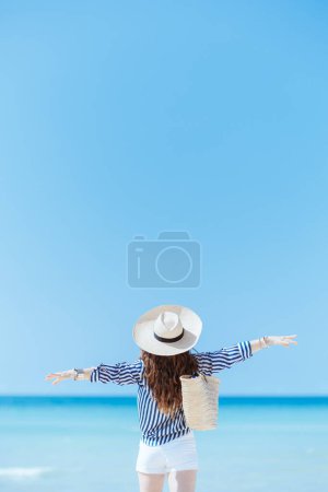 Téléchargez les photos : Vu de derrière femme moderne de 40 ans sur le littoral avec sac de paille et chapeau de paille se réjouissant. - en image libre de droit